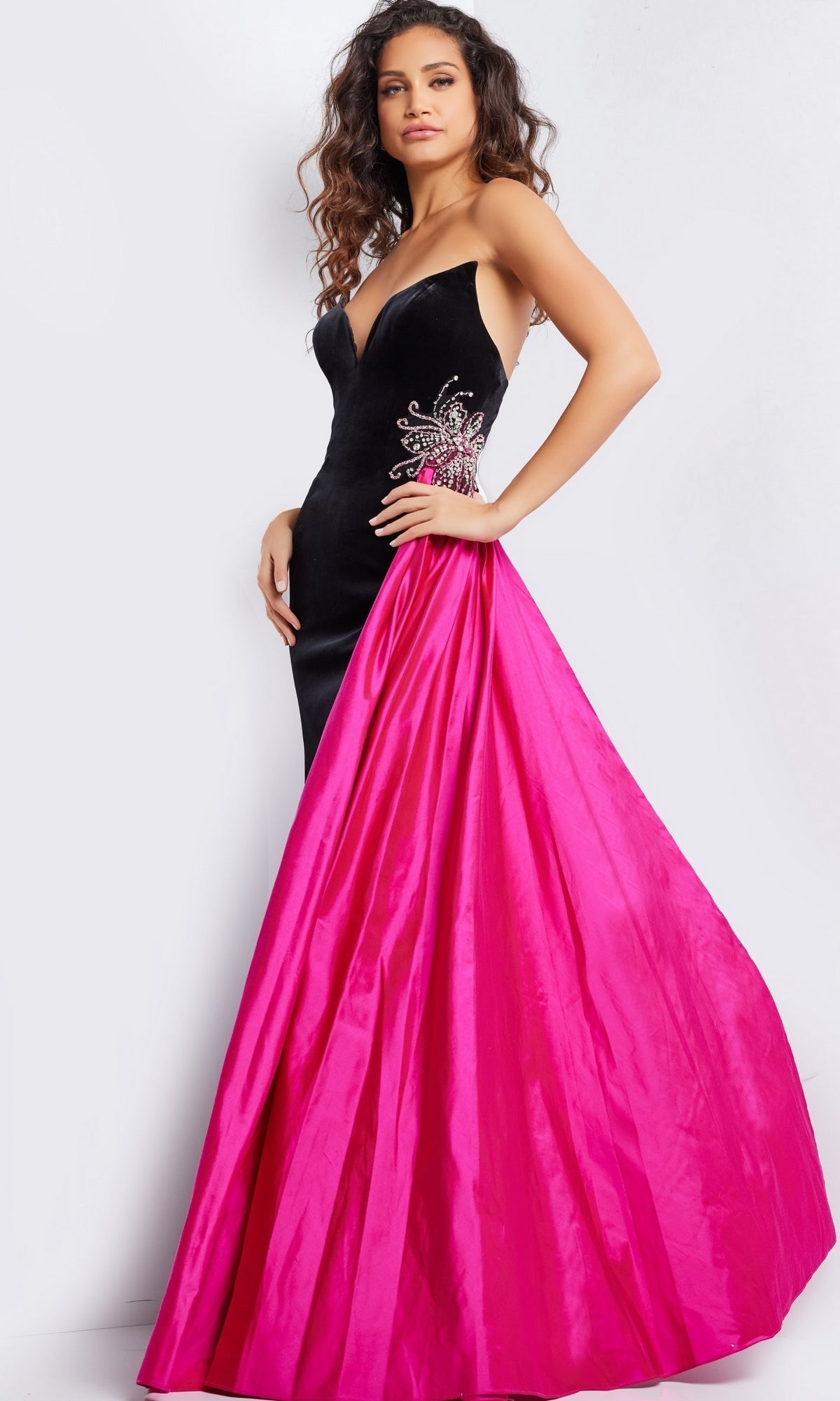 A woman poses in the Jovani Side-Sash Strapless Long Prom Dress 26117, a fitted velvet gown with floral embroidery and a dramatic hot pink satin overskirt—a standout evening look.